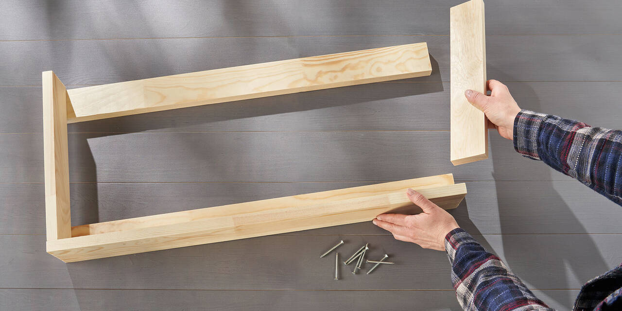 Hände platzieren auf einem Tisch fünf Holzbretter in Form eines Regalrahmens. Daneben liegen Schrauben.